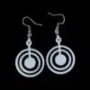 Close-up of white acrylic dangle earrings featuring three concentric circles with a central dot, suspended from silvertone surgical steel ear wires against a black background.