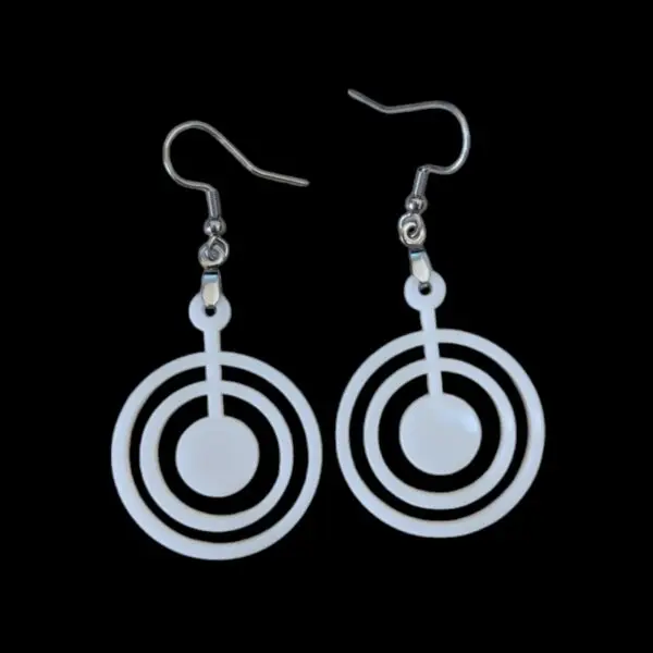 Close-up of white acrylic dangle earrings featuring three concentric circles with a central dot, suspended from silvertone surgical steel ear wires against a black background.