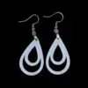 White teardrop-shaped earrings with two nested teardrop cutouts inside, creating a minimalist geometric effect, hanging from silver hooks against a black background.