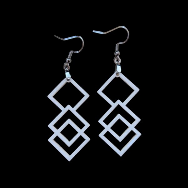 Close-up of white acrylic dangle earrings featuring three interlocking square shapes, suspended from silvertone surgical steel ear wires against a black background.