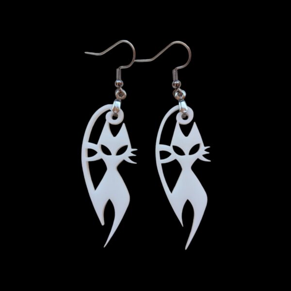 Close-up of white acrylic dangle earrings shaped like stylized cats with pointed ears and whiskers, suspended from silvertone surgical steel ear wires against a black background.