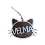 Wooden cat-shaped ornament with pointed ears and whiskers. The name "Velma" is engraved across the body in white text.